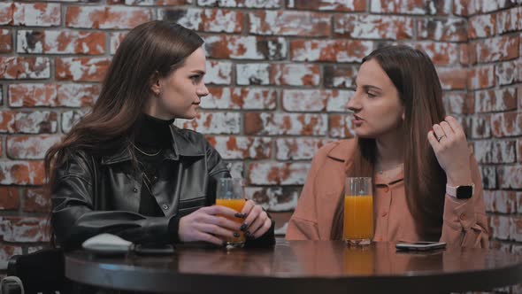 Two Brownhaired Girls Talk in a Cafe Over Glasses of Juice