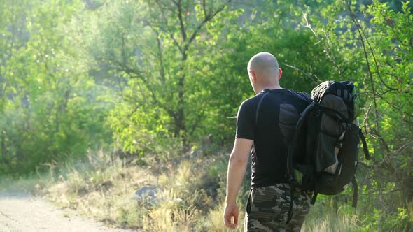 Back view, in movement, a bald hiker man with a backpack on his shoulders walking on a path