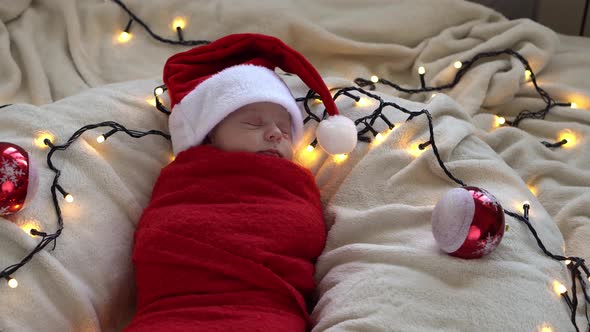 Top View Portrait First Days Of Life Newborn Cute Funny Sleeping Baby In Santa Hat Wrapped In Red