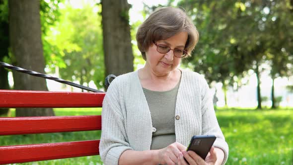 Elderly Woman with a Smartphone in Her Hands Sits on a Bench Surfing the Internet and Researching