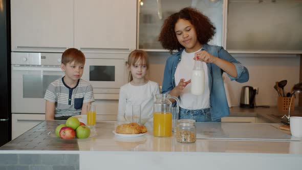 Caring Babysitter Pouring Milk To Boy and Girl Feeding Children in Kitchen at Home