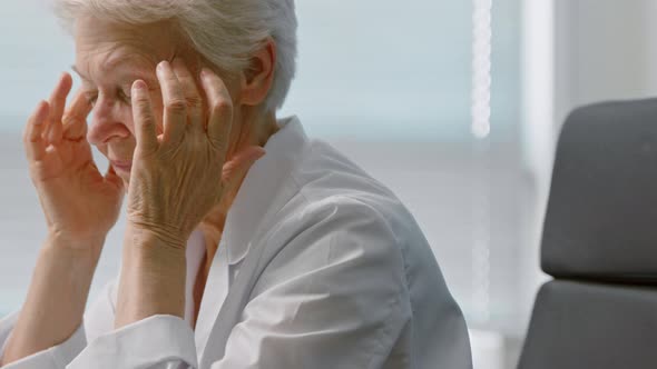 Tired Senior female doctor in coat massages temples suffering from headache