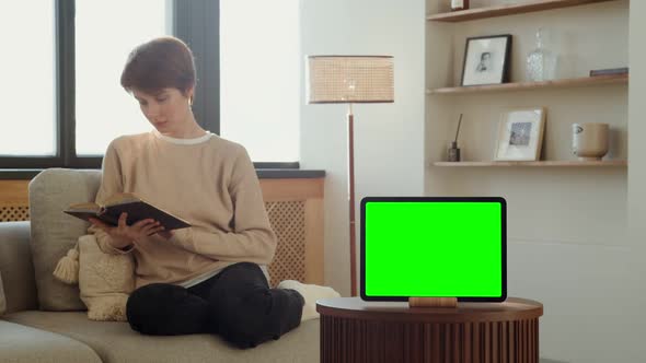 A Tablet with a Green Screen Stands on a Table Against of a Woman Reading a Book