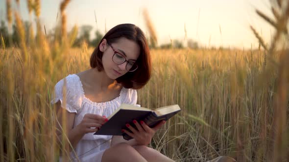 A Young Woman Is Reading a Book in a Yellow Field. A Girl Sits in a Dress in a Wheat Field with a