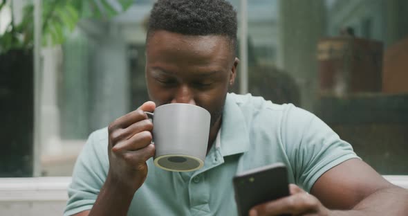 Happy african american man sitting with smartphone and drinking coffee in garden