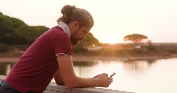 Side View of a Man Using His Gadget While Standing on the Bridge By the Pond