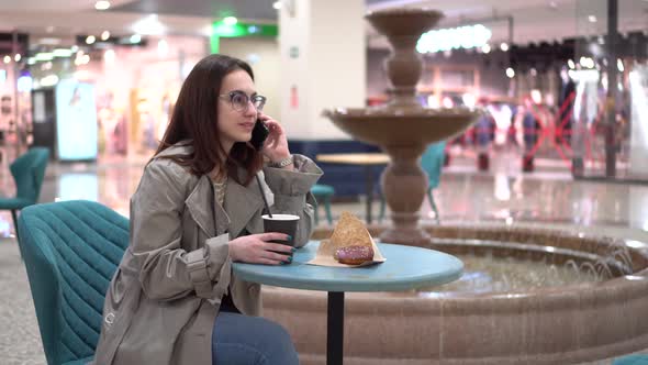 A Young Woman in a Cafe Speaks on the Phone Against the Background of a Fountain