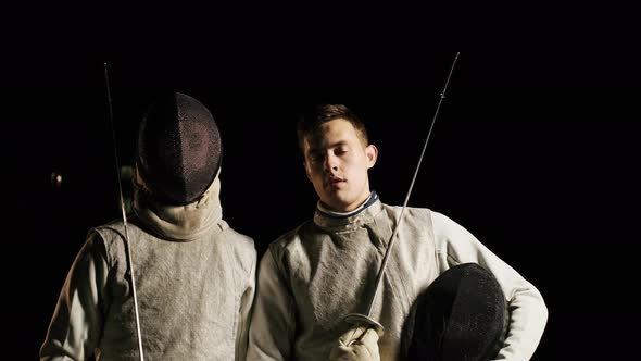 Two Fencers In Suits Stand Next To Each Other In The Dark.