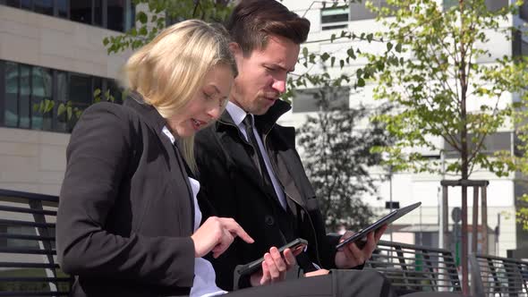 A Businessman and a Businesswoman Sit on a Bench, Work on a Smartphone and a Tablet and Talk