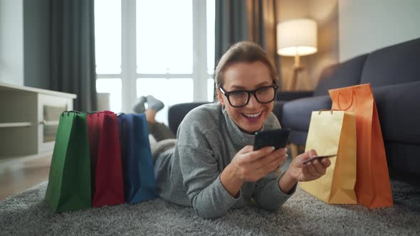 Happy Woman with Glasses Is Lying on the Floor and Makes an Online Purchase Using a Credit Card and
