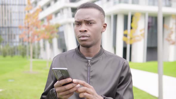 A Young Black Man Works on a Smartphone and Frowns - Office Buildings in the Blurry Background