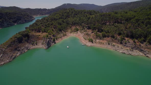 Drone flying to sailship in turquoise water reservoir in Spain with a rocky coast.