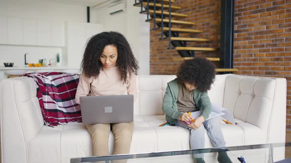 Single Mom Works at Home While Sitting on the Couch While Her Preschooler Son Draws with Felttip