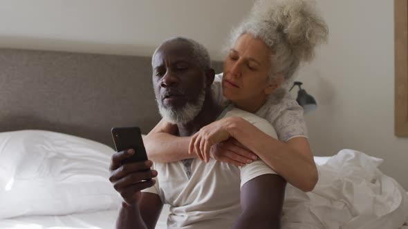 Mixed race senior couple using smartphone while sitting on the bed at home