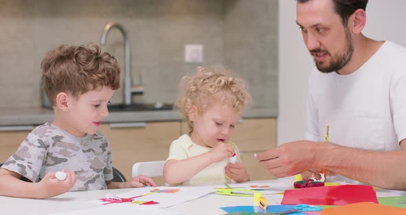 Small Girl and a Small Boy are Making Applications with the Coloured Paper and Glue and Their Father