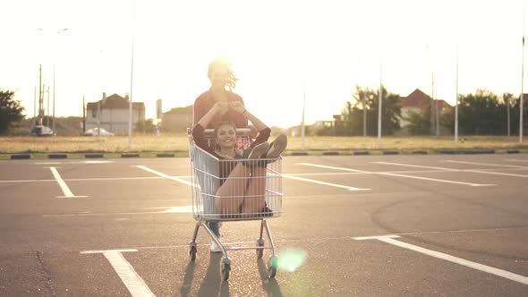 Young Woman is Sitting in the Grocery Cart While Her Friend is Pushing Her Behind in the Parking By