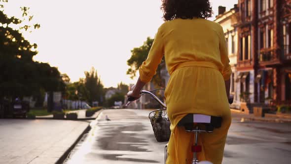 Back View of an Unrecognizable Woman in Long Yellow Dress Riding a City Bicycle with a Basket and