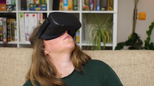 A woman wearing virtual reality glasses sits on a couch and turns her head