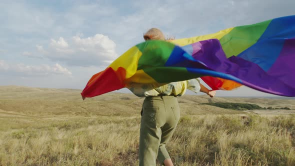 The Girl Is Holding a Large Rainbow Flag and Walking Against the Wind on the Field. View From the