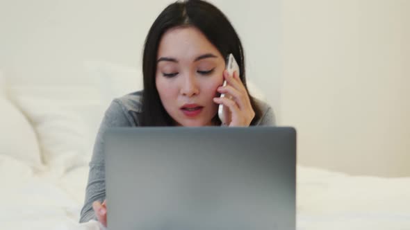 Smiling Asian woman talking by phone and looking at laptop on the bed