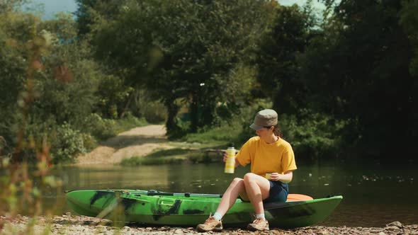 A young woman approaches the kayak and sits down, pouring a drink from a thermos