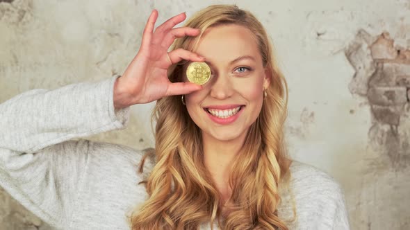 Portrait of Playful Blonde Woman with Charming Smile Standing Covering One Eye with Golden Bitcoin