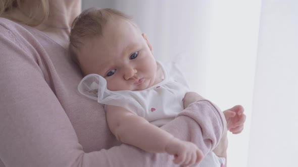 Thoughtful Caucasian Baby Girl in Hands of Unrecognizable Mother at Window Indoors