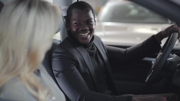 Side View of Blond Caucasian Female Dealer Sitting in Car Salon with Client and Giving Him Car Keys