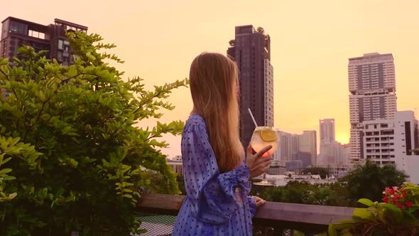 Young Woman in Gown Holding Cocktail on Terrace of Rooftop Bar During Sunset