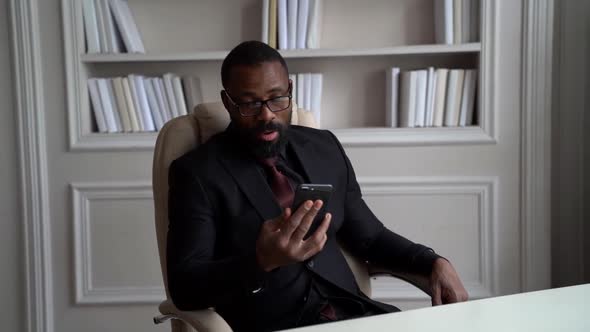 Male Entrepreneur Is Sitting in His Office and Chatting By Video Call in Smartphone