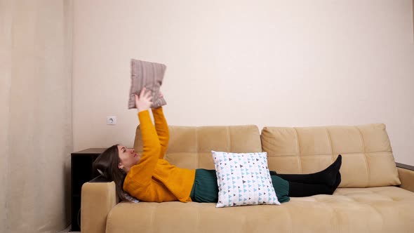 Bored Young Woman in Sweater Lies on Sofa Throwing Pillows