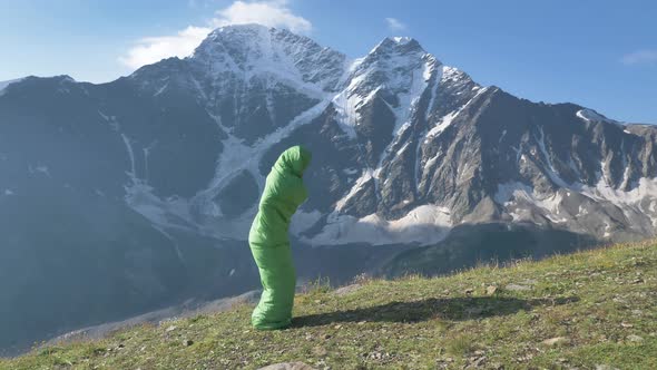Funny Male Traveler Jumps in a Sleeping Bag Against the Backdrop of High Snowy Mountains in Slow