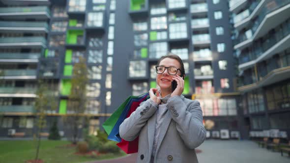 Smiling Woman Walking on a Business District with Shopping Bags and Talking on the Smartphone