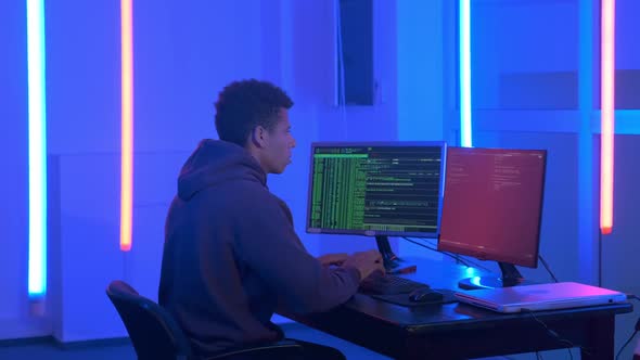 Medium Shot of Young African Man Writing Hacker Programs in Dark Room with Neon Lights