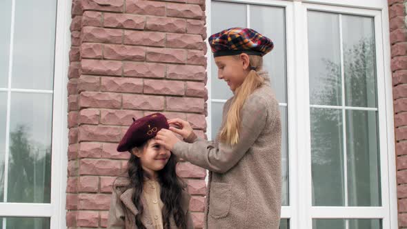 Two Sisters Wearing Beret and Autumn Coat on Brick Wall and Window Background. Older Sister Dressing