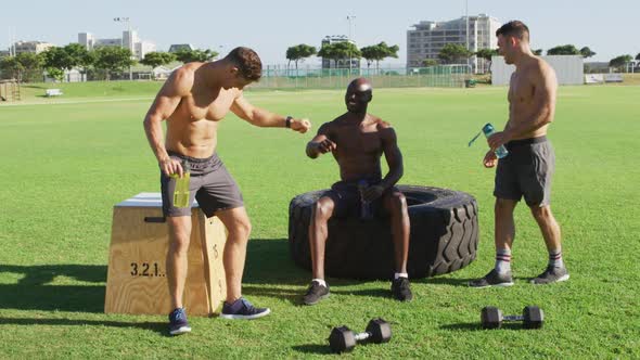 Three fit shirtless diverse men resting, drinking water and talking after exercising outdoors