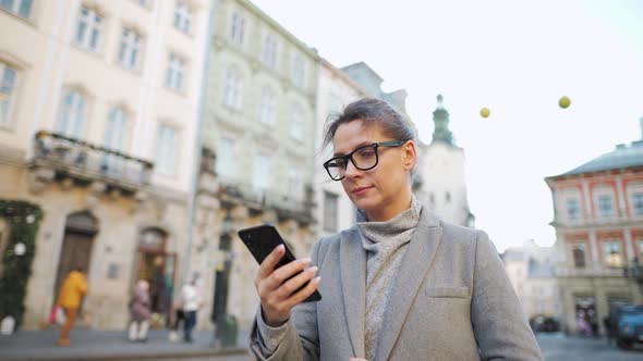 Woman with Glasses Wearing a Coat Walking Down an Old Street and Using Smartphone