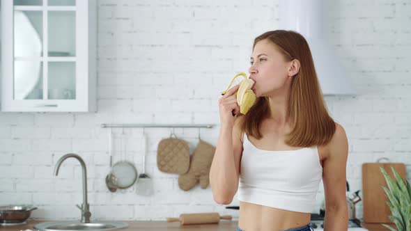A young girl eats a banana on a white kitchen background.