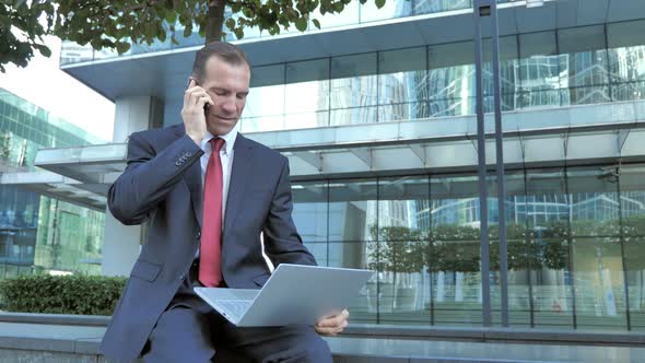 Businessman Talking on Phone While Working on Laptop Outside Office