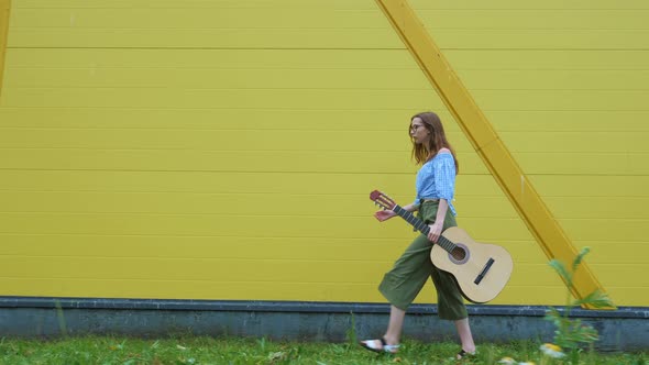 Girl with Guitar Walks Along Yellow Building Wall