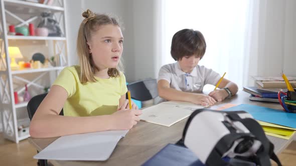 Portrait of Focused Genius Girl Thinking and Writing in Workbook with Blurred Boy Drawing