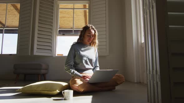 Caucasian woman sitting on floor with cup of coffee using laptop in sunny cottage living room