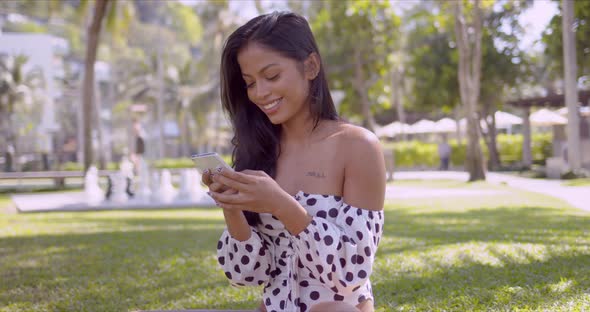 Smiling Asian Woman in Modern Swimsuit Browsing Smartphone Sitting in Park