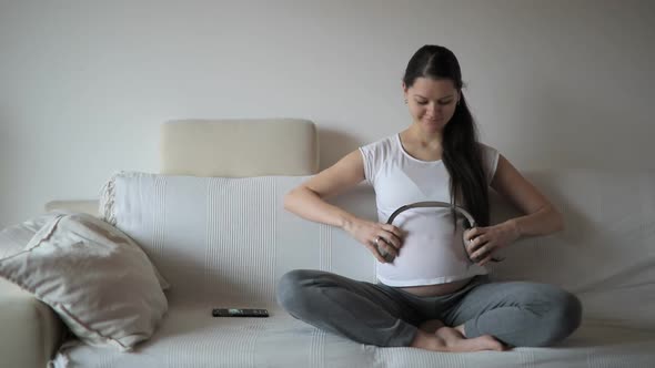 Pregnant Woman Sitting on White Sofa and Keeping Headphones on Belly. Baby Kicks on Music