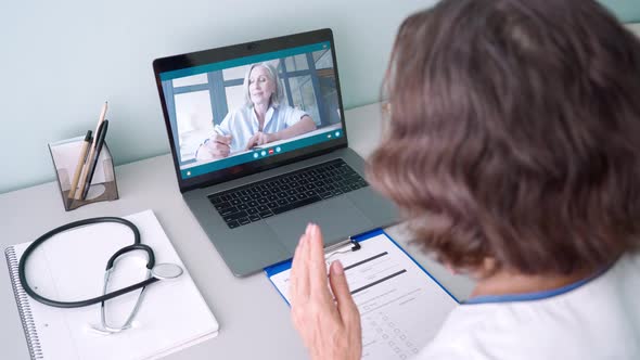 Senior Female Doctor Consulting Remotely Using Laptop Writing Health Data