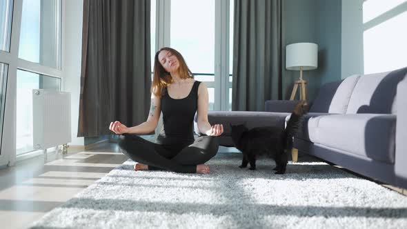 Young Caucasian Woman in Black Jumpsuit Sitting in Lotus Position and Meditating Yoga at Home