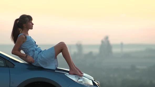 Happy Young Woman Driver in Blue Dress Enjoying Warm Summer Evening Sitting on Her Car Hood