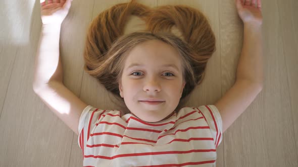 Little Funny Kid Lying on the Floor and Looking Into Camera at Room. Happy Smiling Blonde Girl