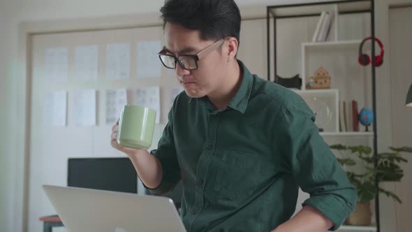 Designer Sits On His Desk Holds Laptop On The Knees Drinks Coffee While Working On The Project