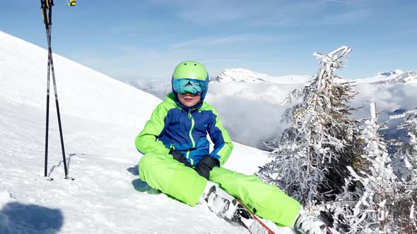 Smiling Boy with Alpine Ski and Helmet Sit in Snow Lifting Hands